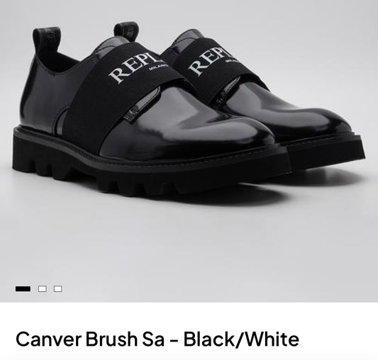 A pair of black patent leather slip-on shoes with wide black elastic straps featuring the white 'REPLAY' logo, set against a light gray background.