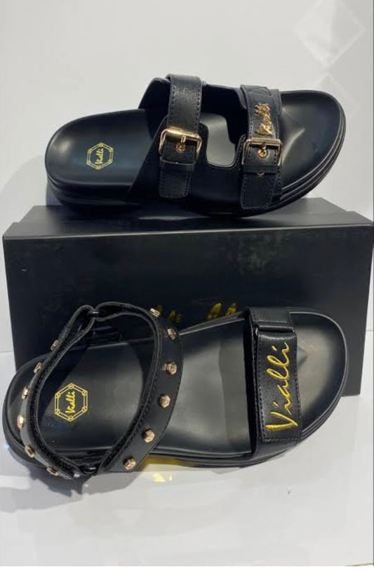 Black double-strap sandals with gold-tone buckles and studs, featuring the Vialli brand name in gold on the strap, displayed on a black shoebox.